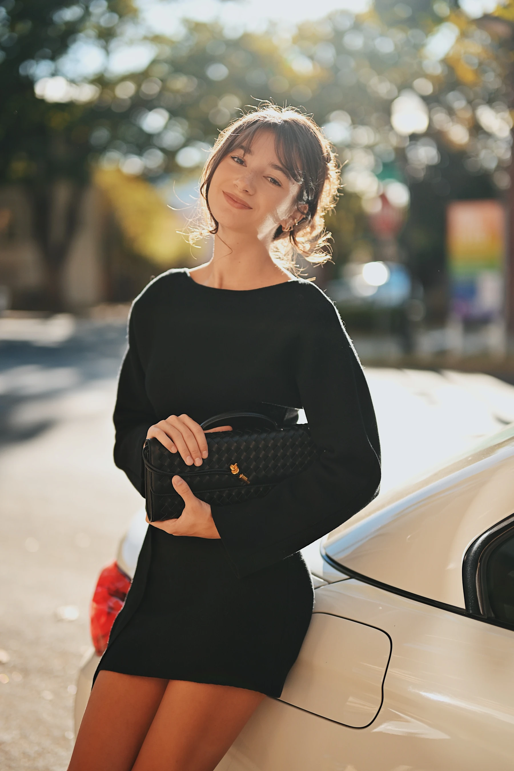 Rosina leaning on a cream white BMW holding a woven clutch, backlit hair